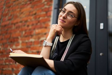 Gündüz gözlüklü genç beyaz kadın portresi şehirde öğrenim ve öğrenim görmek için kameraya bakıyor. Eğitim çizim defteriyle dışarıda poz veren çekici bir kız öğrenci.