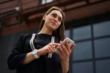 Modern mobil teknolojiyi elinde tutan ve sosyal medyaya yayın paylaşımını düşünen, dijital akıllı telefonlu kadın blogcu şehir caddesinde hayal kuruyor.