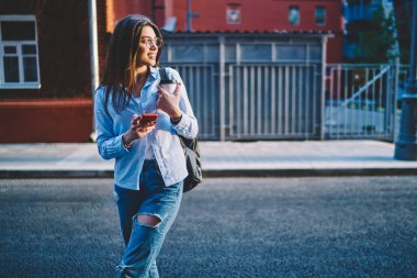 Güneşli seyahat gününde boş zamanlarında dijital akıllı telefonlu çekici genç bir kadın, gözlüklü neşeli hippi kız cep telefonu ve 4G bağlantısı kullanarak turistik caddeden uzağa bakıyor.