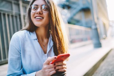 Görüntü koruması için şık gözlükleri olan genç bir kadın elinde blogla gülümseyen bir akıllı telefon, modern cep telefonlu beyaz bir kız boş zamanlarının ve güneşli bir günün tadını çıkarıyor.