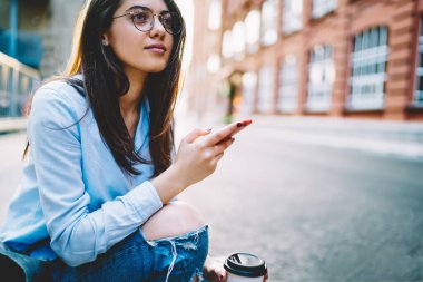 Optik gözlüklü düşünceli bir kadın akıllı telefonu elinde tutuyor ve internette sohbet etmeyi düşünüyor. Şehir merkezinde cep telefonu kullanan düşünceli bir hippi kız. Ağ metinleri üzerinde kafa yoruyor.