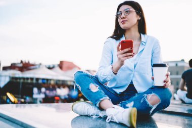 Düşünceli, gündelik giyinmiş genç bir turist elinde kahve, elinde cep telefonu seyahat sırasında düşünceli, düşünceli, cep telefonlu bir blogcu.