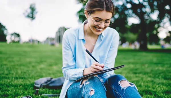 緑の芝生の上でスケッチブックを持って屋外で仕事を楽しんでいる陽気な女性デザイナー ノートに描いた幸せな才能のある女性 — ストック写真