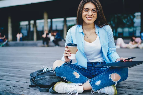 穿着光学眼镜 拿着咖啡去的快乐的年轻女子的画像 拿着素描本和咖啡因饮料 站在室外摆出荷花姿势的相册作画 — 图库照片