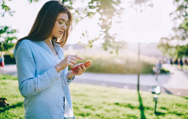アイウェアの若いヒップスターの女の子は スマートフォンのプレイリストに曲をダウンロード春の日を過ごす公園で レジャーで携帯電話やイヤフォンを使用して無線アプリケーションに接続する女性の千年紀 — ストック写真