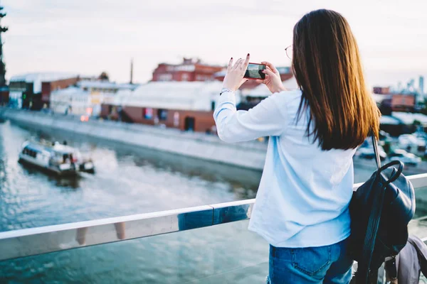 スマートフォンのデバイス上で川の写真を作る上で モバイルカメラに焦点を当てた100万人の女性観光客の背面ビュー カジュアル旅行ブログに共有するための写真をクリックしてバックパックでヒップスターの女の子を身に着けて — ストック写真