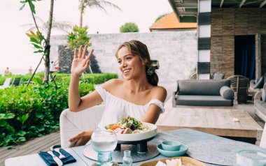 İyimser kadın gülümseyip el sallarken yüzünü çeviriyor ve açık hava restoranındaki masada oturmuş taze sağlıklı salatanın tadını çıkarıyor.
