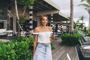 Bulutlu bir günde tropikal kafe terasında dururken cep telefonu kullanıp gülümseyen yaz kıyafetli mutlu genç bir bayan.