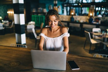 Yukardan gülümseyen beyaz bluzlu bir bayan modern netbook 'ta internette sörf yaparken rahat bir restoranda akıllı telefonuyla oturuyor.