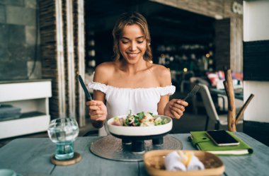 Smiling stylish young female in trendy top with open shoulders sitting at table in cozy spacious cafe with cutleries and enjoying tasty vegetable salad served in creative bowl clipart