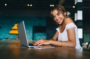 Modern çalışma alanında yüksek masada oturup gülümserken modern taşınabilir netbook üzerinde çalışan şık kıyafetli arkadaş canlısı genç bayan.