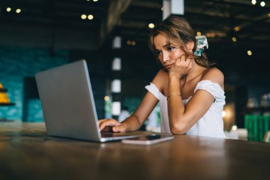 Modern netbook kullanan, şık beyaz bluzlu hoşnutsuz genç bir kadın yaratıcı iş yerinde masada otururken ve el ele tutuşurken.