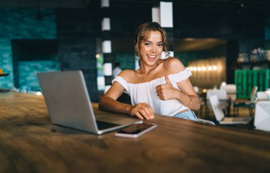İyimser gülümseyen, modaya uygun beyaz bluzlu genç bayan, restoranda çağdaş netbook ve akıllı telefonuyla masada oturmuş kameraya bakıyor ve işaret dilini gösteriyor.