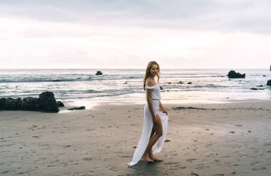 Okyanusun kıyısındaki kumlu sahilde duran ve yaz gününün tadını çıkaran beyaz maksi saçlı, mutlu, genç, etnik bir bayan turist.