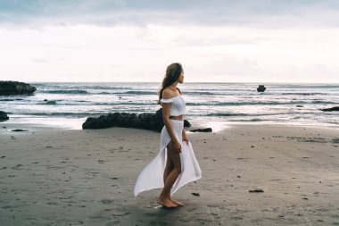 Beyaz elbiseli, bronz tenli genç bir kadın gezginin boş kumsalda tek başına durup yaz tatili boyunca bakışlarını kaçırması.