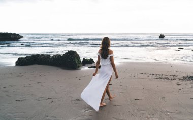 Plajda yürüyen ve tropikal bir ülkede yaz tatili boyunca okyanusa el sallayan beyaz maksimumlu çıplak ayaklı genç bayanın arka planda görüntüsü.