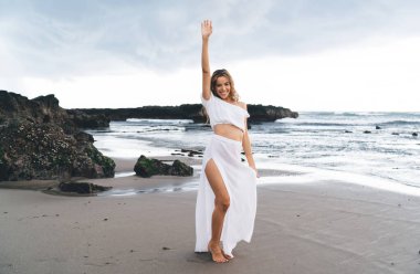 Kolunu kaldırmış, beyaz ışıklı bir elbiseyle kayalık okyanus kıyısındaki kumlu sahilde duran ve yaz tatilinin tadını çıkaran heyecanlı genç bir bayan.