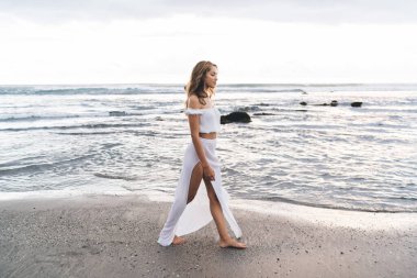 Beyaz bluzlu kahverengi saçlı, çıplak ayaklı genç bir kadın rüzgarsız bir günde kumsalda geziniyor.