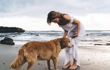 Beyaz elbiseli neşeli bir kadın kumlu sahilde duruyor ve tatilde Golden Retriever köpeğini okşuyor.
