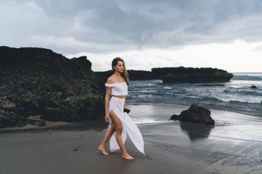 Beyaz elbiseli çıplak ayaklı, dalgın bir kadın kumsalda yürüyor, deniz kenarında ve tatildeyken başka tarafa bakıyor.