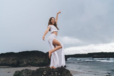 Kayalık sahilde duran, gülümseyen ve kameraya bakarken poz veren beyaz, ince, havalı elbiseli bir kadın.