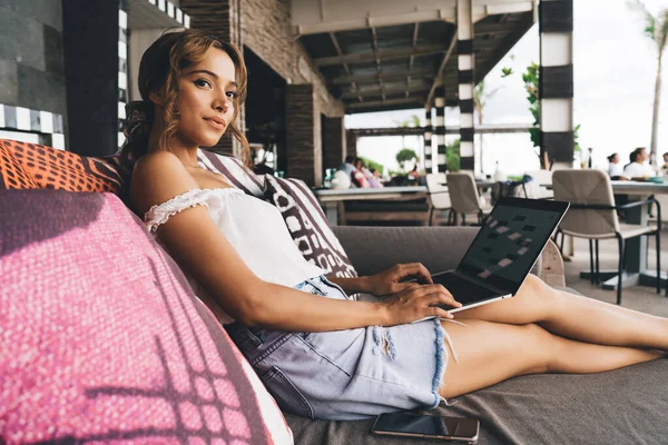 夏の日に現代ホテルのベランダでソファにくつろぎながらノートパソコンを閲覧するカジュアルな服の若い女性の肯定的な側面図 — ストック写真