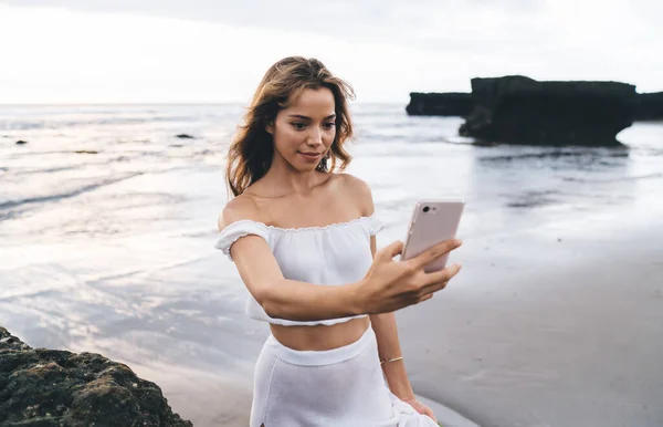 年轻的心满意足的女性 棕色头发 头顶白皙 肩膀张开 裙子敞开 在手机上对着大海自拍 — 图库照片