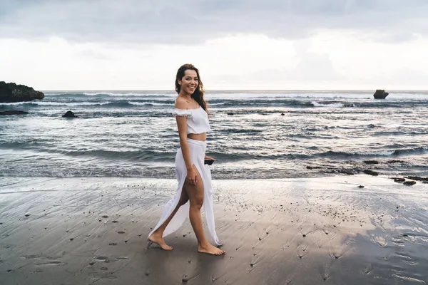 Beyaz giyen zarif, çıplak ayaklı kadın kumlu sahilde durup kameraya bakarken gülümsüyor.