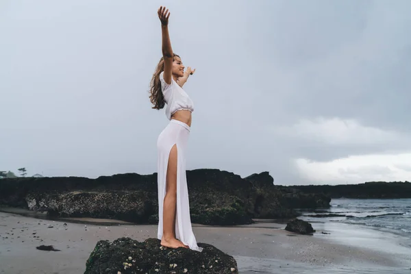 腕を上げ 自由を楽しんで岩の崖の上に立って白いドレスの完全な長さ幸せな裸足の女性の側面図 — ストック写真