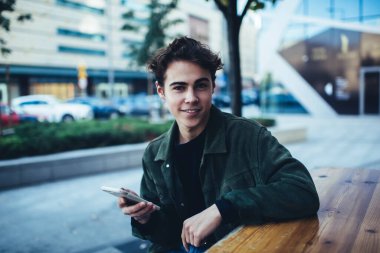 Sokakta masada oturmuş cep telefonuyla sohbet ederken günlük kıyafetleriyle kameraya bakan genç ve mutlu bir erkek.