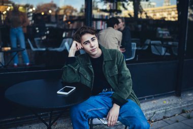 Gündelik kıyafetli genç bir erkek öğrenci. Yuvarlak masada oturmuş kameraya bakıyor. Sırtüstü uzanıyor ve ara veriyor.