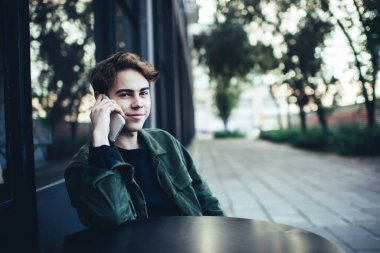 Genç, mutlu erkek, günlük giysiler giyip kameraya bakarken akıllı telefon kullanarak caddedeki kafede otururken arama yapıyor.