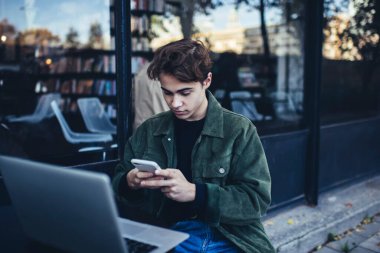 Ciddi bir erkek öğrenci günlük giysiler içinde sokakta masada oturuyor ve ödev yaparken cep telefonuyla internette geziniyor.