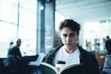 Açık kafede üniversite ödevi yaparken kameraya bakan düşünceli erkek öğrenci kahve servisi ve ders kitabıyla oturuyor.