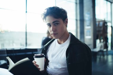 Düşünceli bir erkek öğrenci, kafeteryada oturmuş ödevlerini yaparken elinde bir fincan kahveyle notlar okurken kameraya bakıyor.