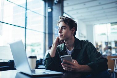 Düşünceli, uzaktan kumandalı bir erkek, elinde dizüstü bilgisayarla masada oturuyor ve iş için cep telefonunu kullanıyor.