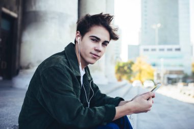 Sokak ortasında oturmuş müzik dinlerken kameraya bakan ve akıllı telefon kullanan düşünceli bir erkeğin yan görüntüsü.