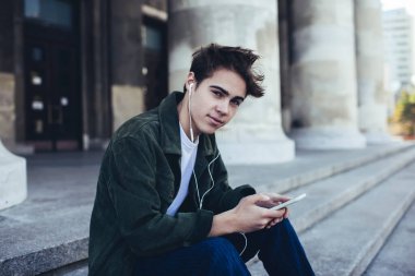 Gülümseyen erkek sokakta oturmuş kulaklıkla müzik dinlerken ve molada cep telefonuyla sohbet ederken kameraya bakıyor.