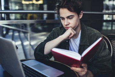Kafede oturan, elinde not defteri olan ve çalışırken laptoptan bilgi okuyan, günlük kıyafetli, odaklanmış bir erkek uzaktan çalışan.
