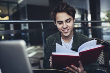 Zevkli erkek öğrenci dişlek bir gülümsemeyle masada oturmuş sınav için hazırlanırken notları okuyor.