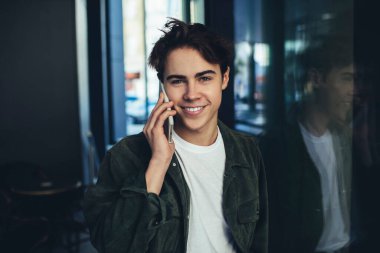 Cam duvara yaslanmış ve cep telefonuyla konuşurken kameraya bakan dişlek, iyimser genç bir erkek portresi.
