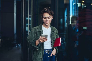 Elleri cebinde, planlamacı ve modern odada cep telefonuyla mesajlaşan iyimser genç bir erkek.