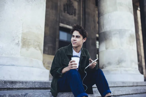コーヒーを飲みながら階段に座り イヤホンで音楽を聴きながら笑顔で微笑む陽気な男性 — ストック写真