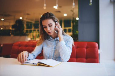 Genç, konsantre bir kadın, kafeteryada cep telefonuyla konuşurken açık not defteriyle oturup notlar okuyor.