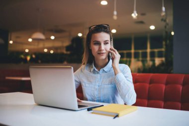 Sakin kadın serbest çalışan, günlük kıyafetlerini giymiş, hafif çalışma odasında dizüstü bilgisayarla otururken yüzünü çeviren ve aramak için akıllı telefonu kullanan biri.