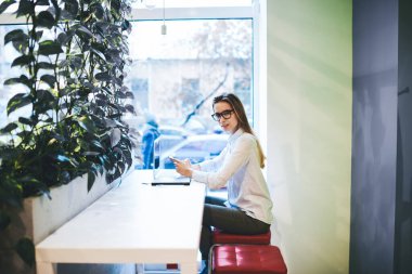 Günlük giysiler içinde, netbook ile masada oturan ve gündüz kafede akıllı telefondan mesajlaşan pozitif kız öğrencilerin yan görüntüsü.