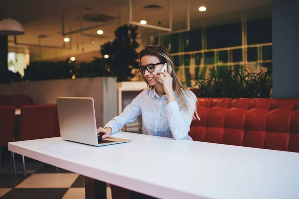 Mujer Sonriente Freelancer Anteojos Camisa Ligera Escribiendo Laptop Hablando Smartphone —  Fotos de Stock