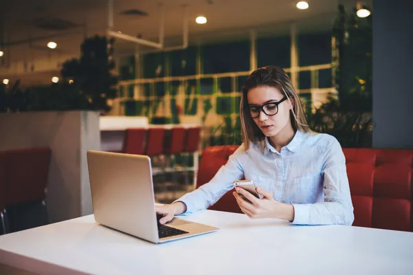 有思想的女自由职业者 白天在现代咖啡馆工作时 戴着眼镜 穿着浅色衬衫 一边浏览笔记本电脑 一边用智能手机 — 图库照片