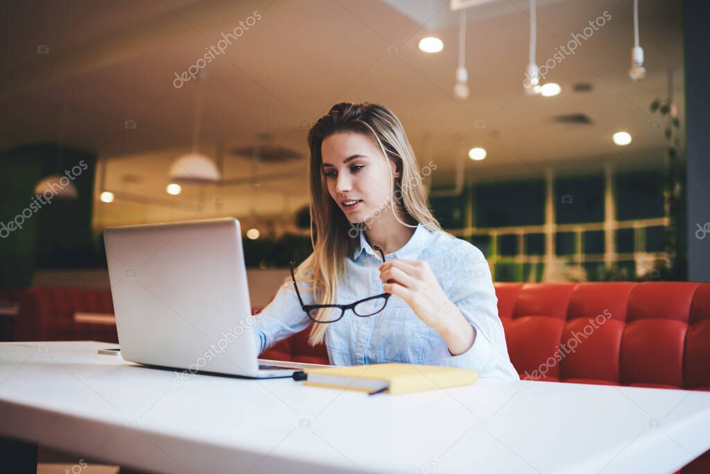 Pensive female freelancer in casual outfit with eyewear sitting at table in light cafeteria and using laptop while doing work