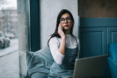 Düşünceli kadın uzaktan kumandalı, günlük giysiler içinde dizüstü bilgisayarla otururken ve çalışırken akıllı telefondan konuşurken başka tarafa bakıyor.